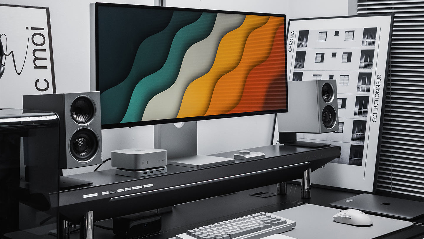 A pair of black SOVOX MiniMax3 Monitor speakers on a desk shelf either side of an ultrawide display